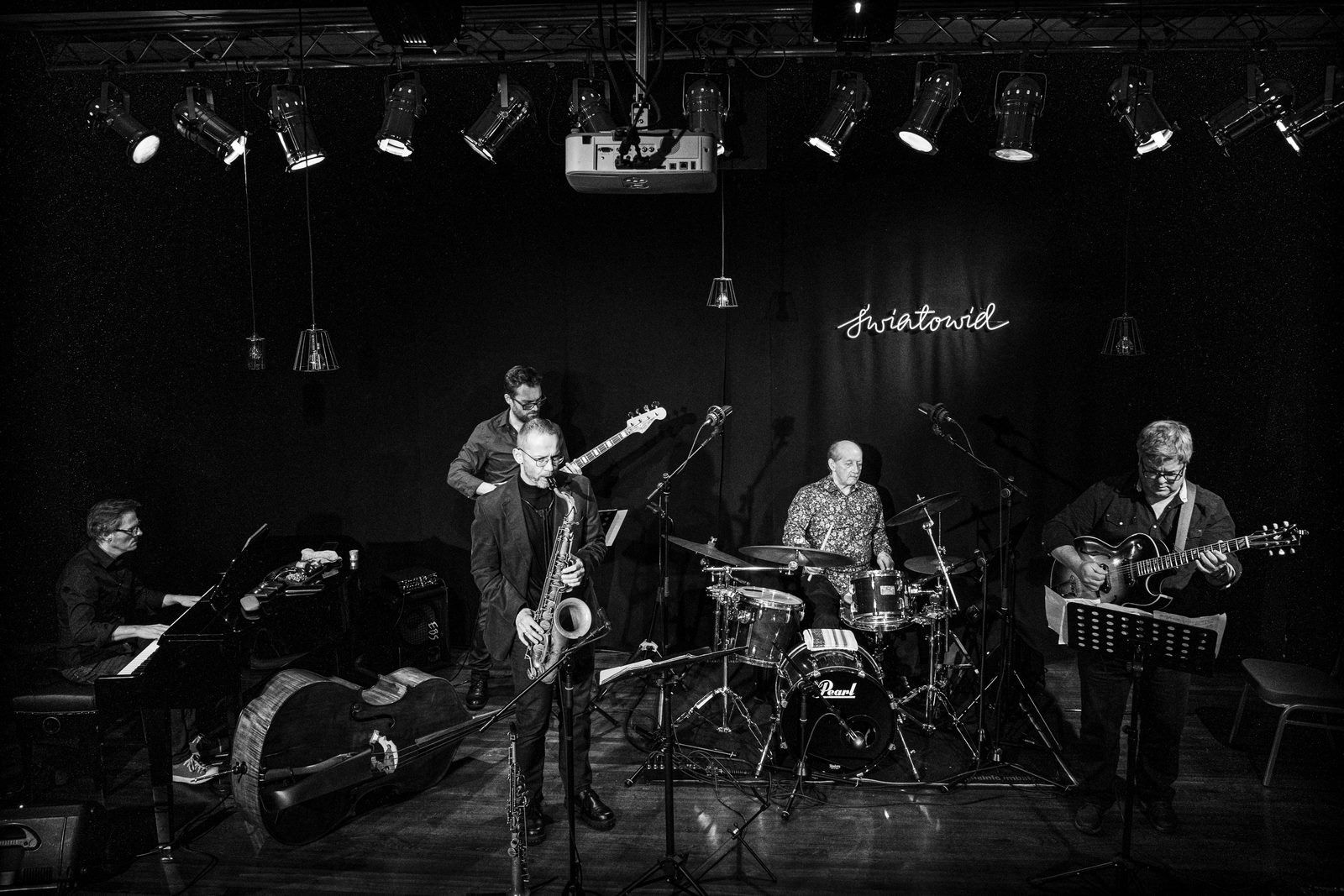 Zdjęcie czarno-białe; na scenie pianista, saksofonista, basista i gitarzysta ubrani na ciemno, perkusista we wzorzystej koszuli