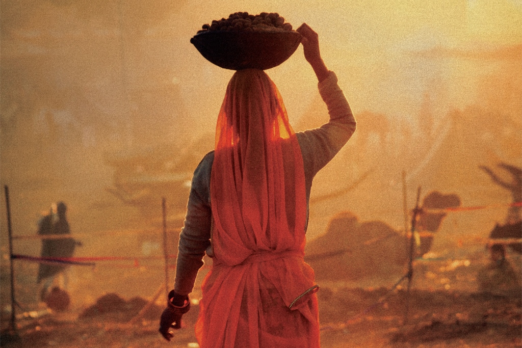 Hinduska w długiej szacie i czerwonym przezroczystym welonie stoi tyłem przytrzymując ręką kosz, który ma na głowie