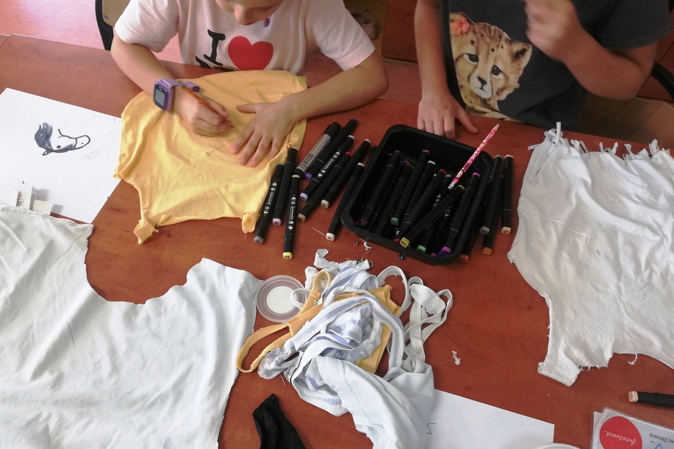 Stół warsztatowy, rozłożone koszulki trykotowe, z których uczestnicy wycinają różne kształty