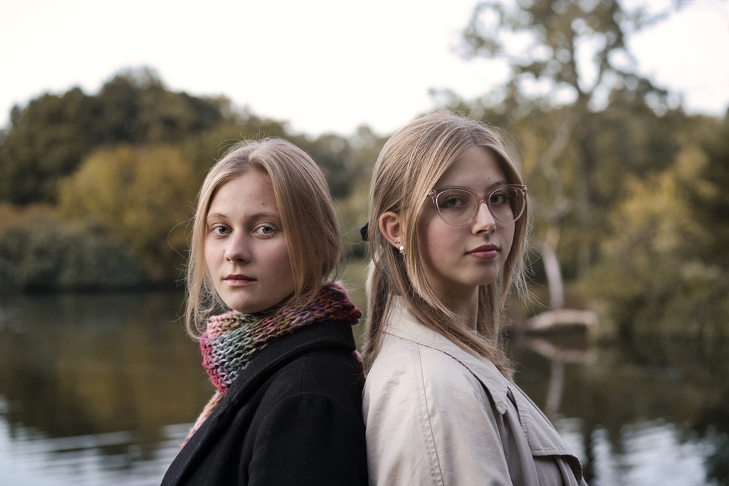 Dwie dziewczyny, blondynki ze spiętymi do tyłu włosami, jedna w jasnej druga w czarnej marynarce stoją zwrócone plecami do siebie nad rzeką