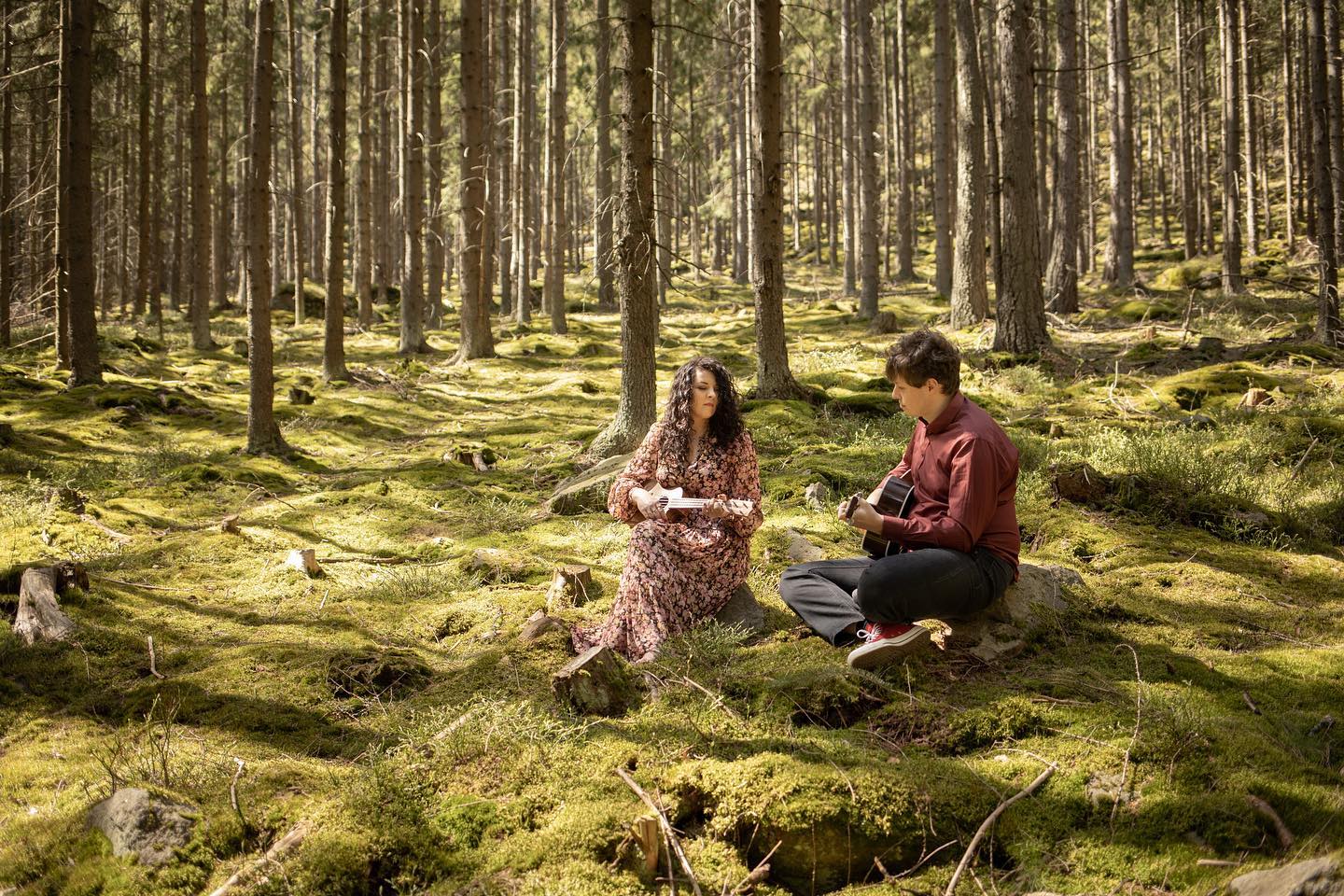 w lesie na mchu siedzą chłopak z gitarą i dziewczyna z długimi kręconymi włosami trzymająca ukulele