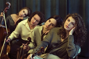 Kobiecy kwartet smyczkowy, wiolonczelistka i alcistka siedzą na ozdobnej sofie trzymając instrumenty przed sobą, obie skrzypaczki stoją za nimi