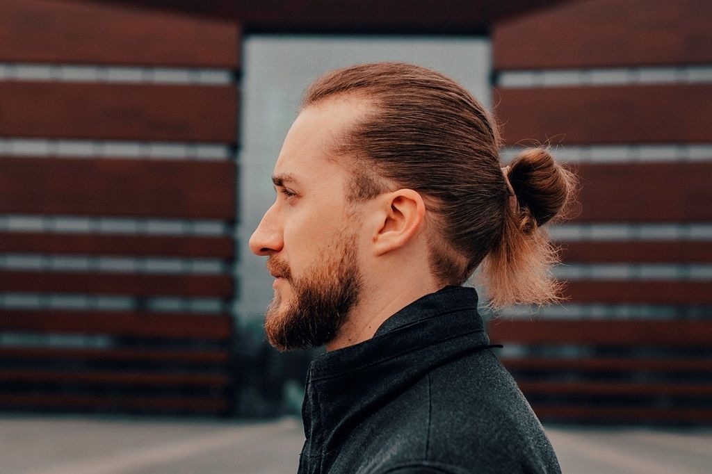 profil mężczyzny, z krótką brodą, włosy związane w węzeł, ciemne ubranie, na tle budynku Narodowego Forum Muzyki