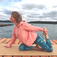Kobieta z kręconymi włosami w jednej z pozycji jogi siedzi na dywaniku rozłożonym na pomoście nad jeziorem.