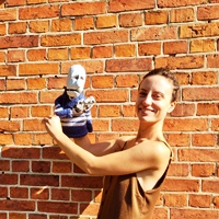 Uśmiechnięta dziewczyna na tle ceglanego muru, w wyciągnietych rękach trzyma teatralną prostą lalkę