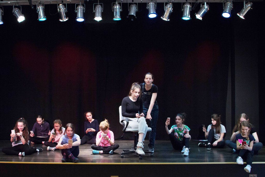 zdjęcie przedstawia fragment spektaklu -  grupę dzieci siedzącą na scenie i zapatrzoną w swoje telefony komórkowe. Pomiędzy nimi na krześle siedzi dziewczyna, także patrzy na swój telefon, a osoba stojąca za nią też zerka na ekran
